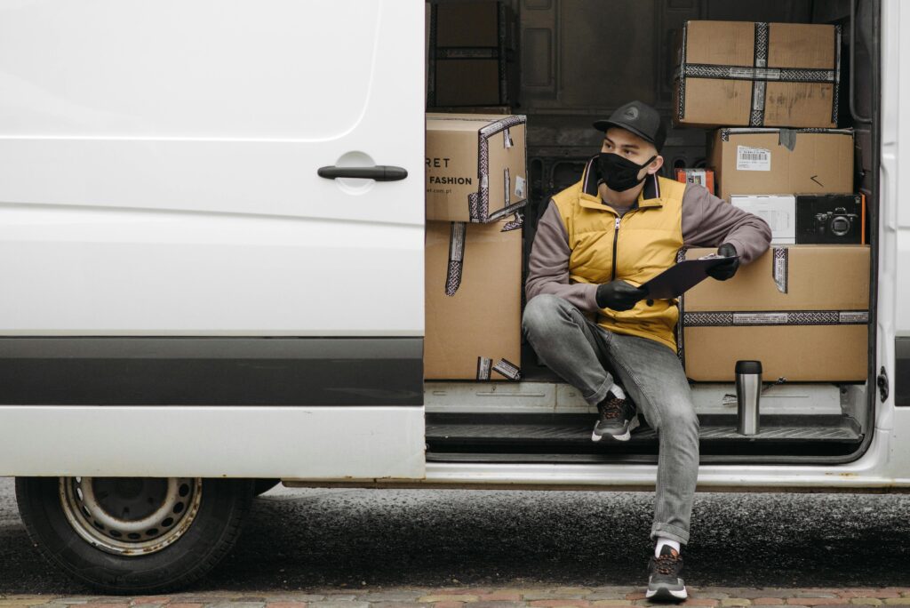 pexels-photo-6407524-6407524 Courier with mask holding clipboard, sitting in delivery van loaded with packages.